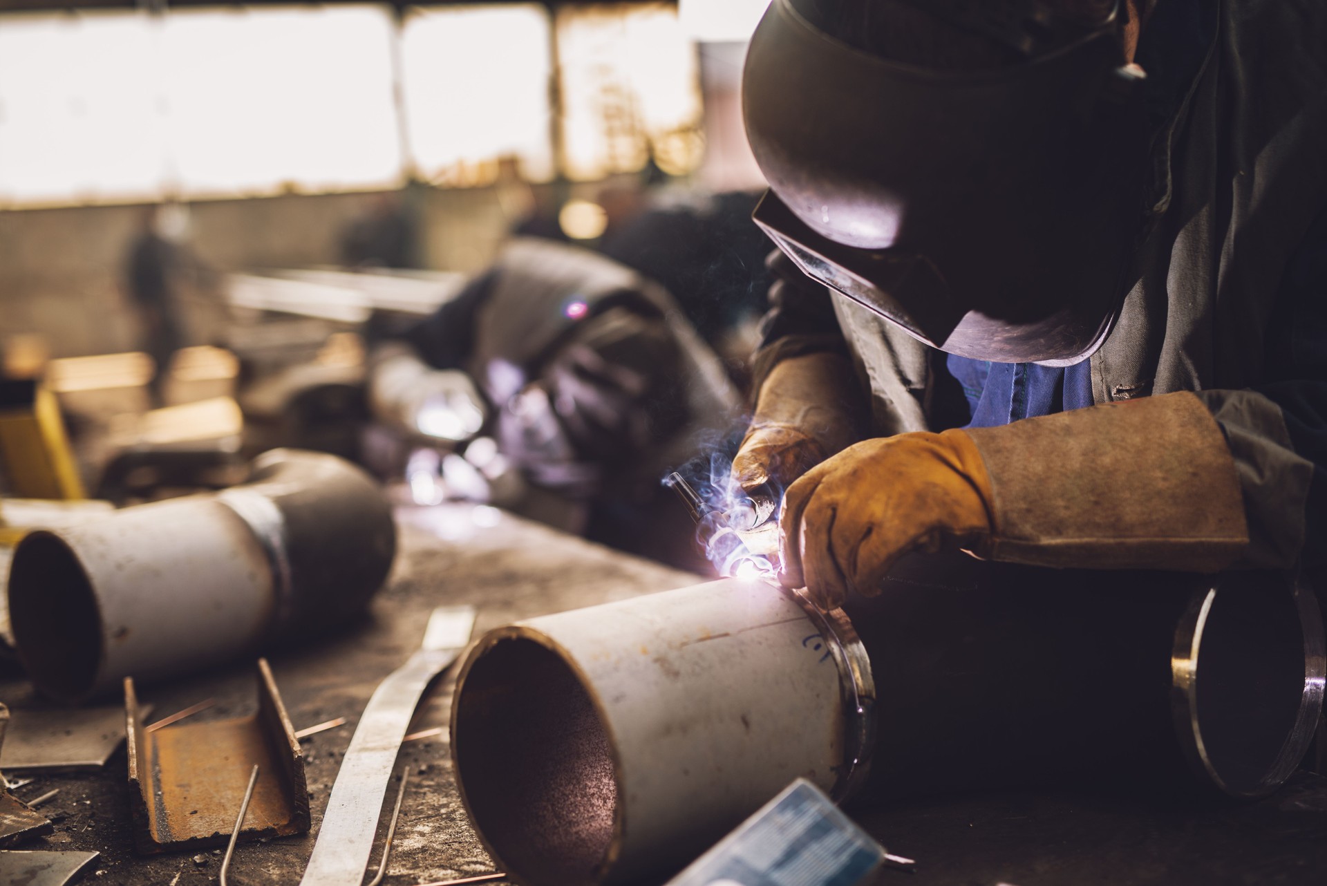 Welder in protective uniform and mask welding metal pipe on the industrial table while sparks flying.